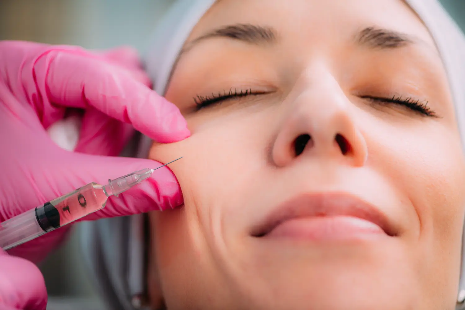 Close-up of a syringe being injected into a person’s cheek while a practitioner in pink gloves supports the skin.