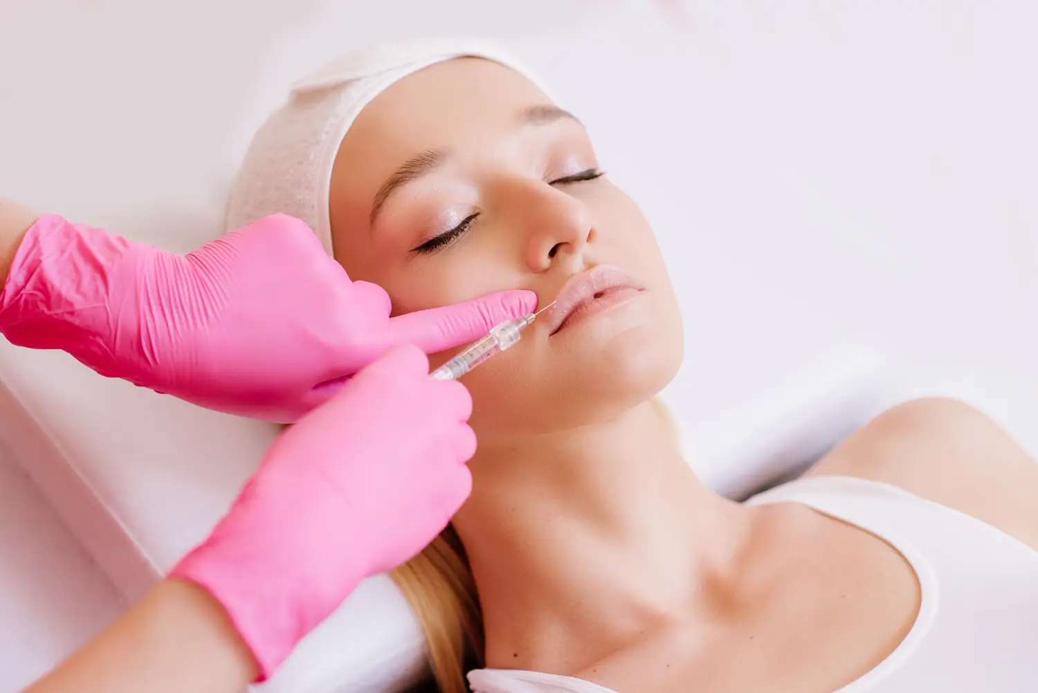Practitioner wearing pink gloves injects a syringe into the upper lip of a person lying on a treatment chair.