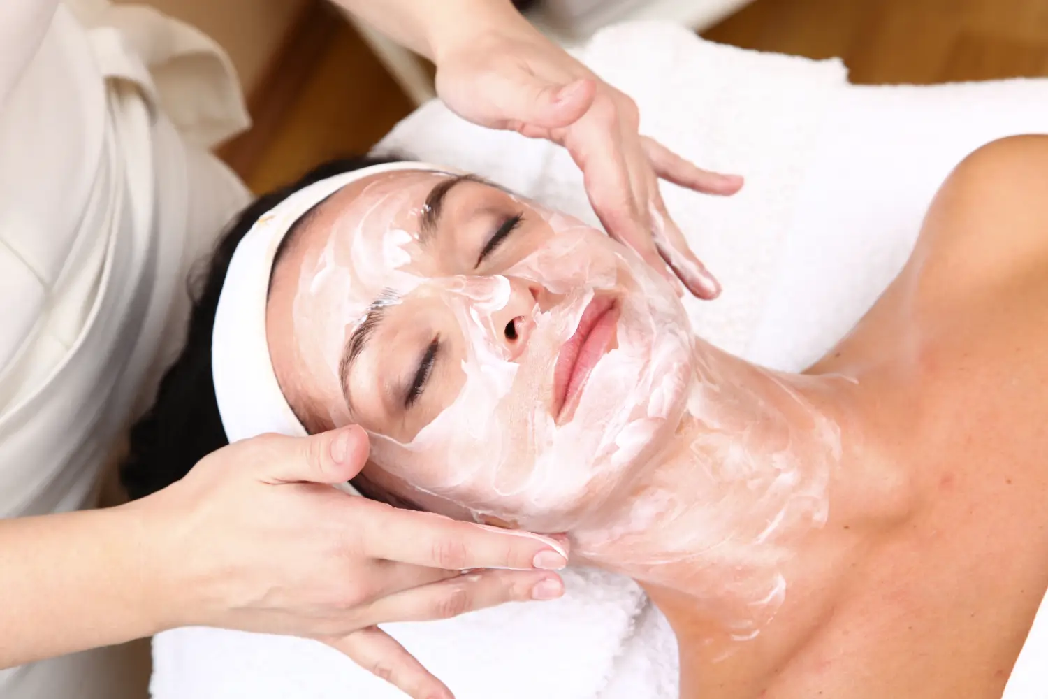 Person lying on a spa bed with eyes closed while hands apply white cream across the face and neck.