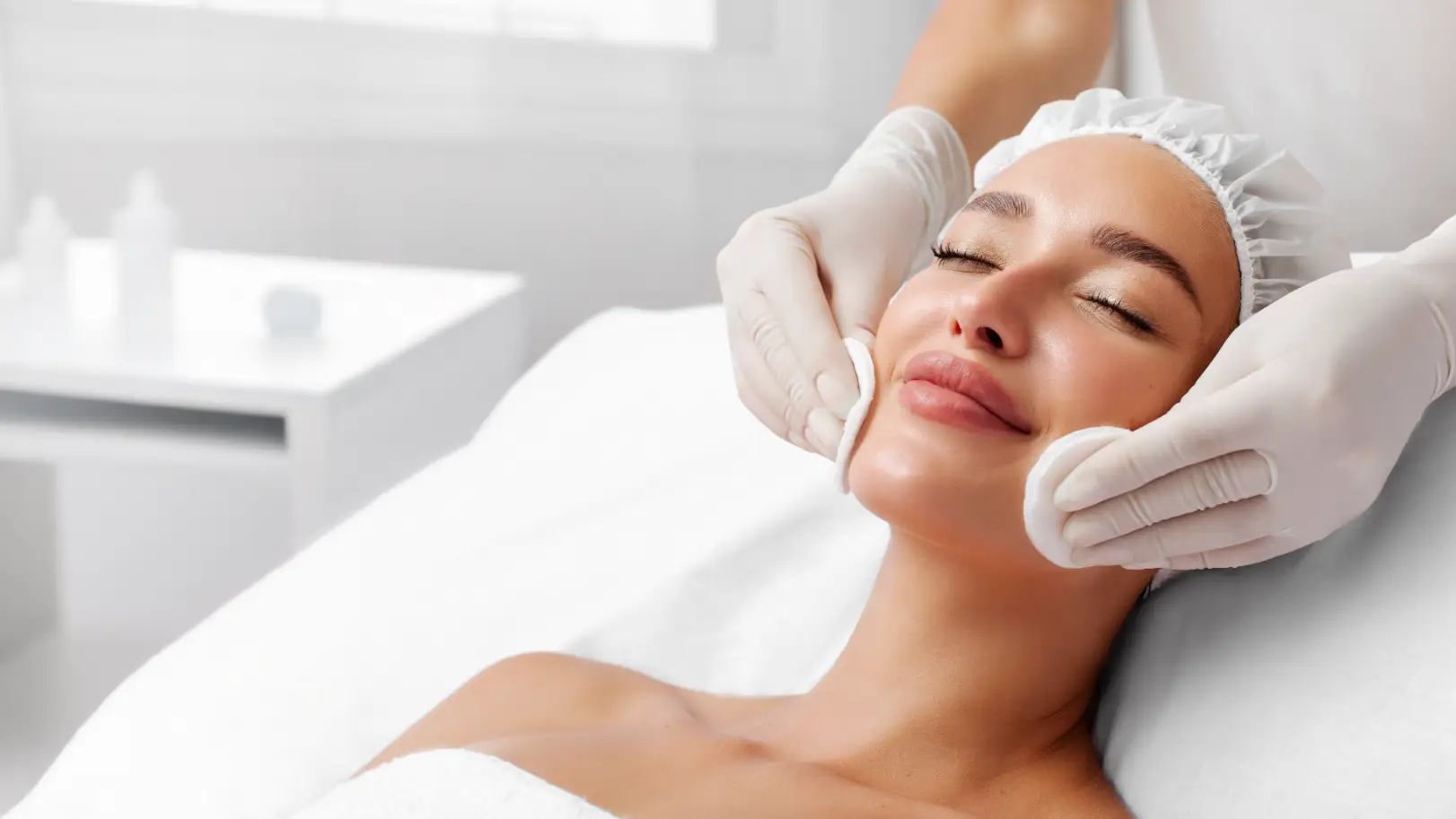Person reclining on a treatment bed while gloved hands cleanse the face with cotton pads.