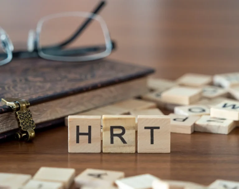 Wooden blocks spelling HRT beside a book and eyeglasses on a table.