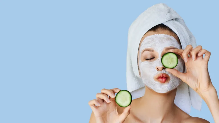 Person with a facial mask and towel holds cucumber slices near the face against a blue background.