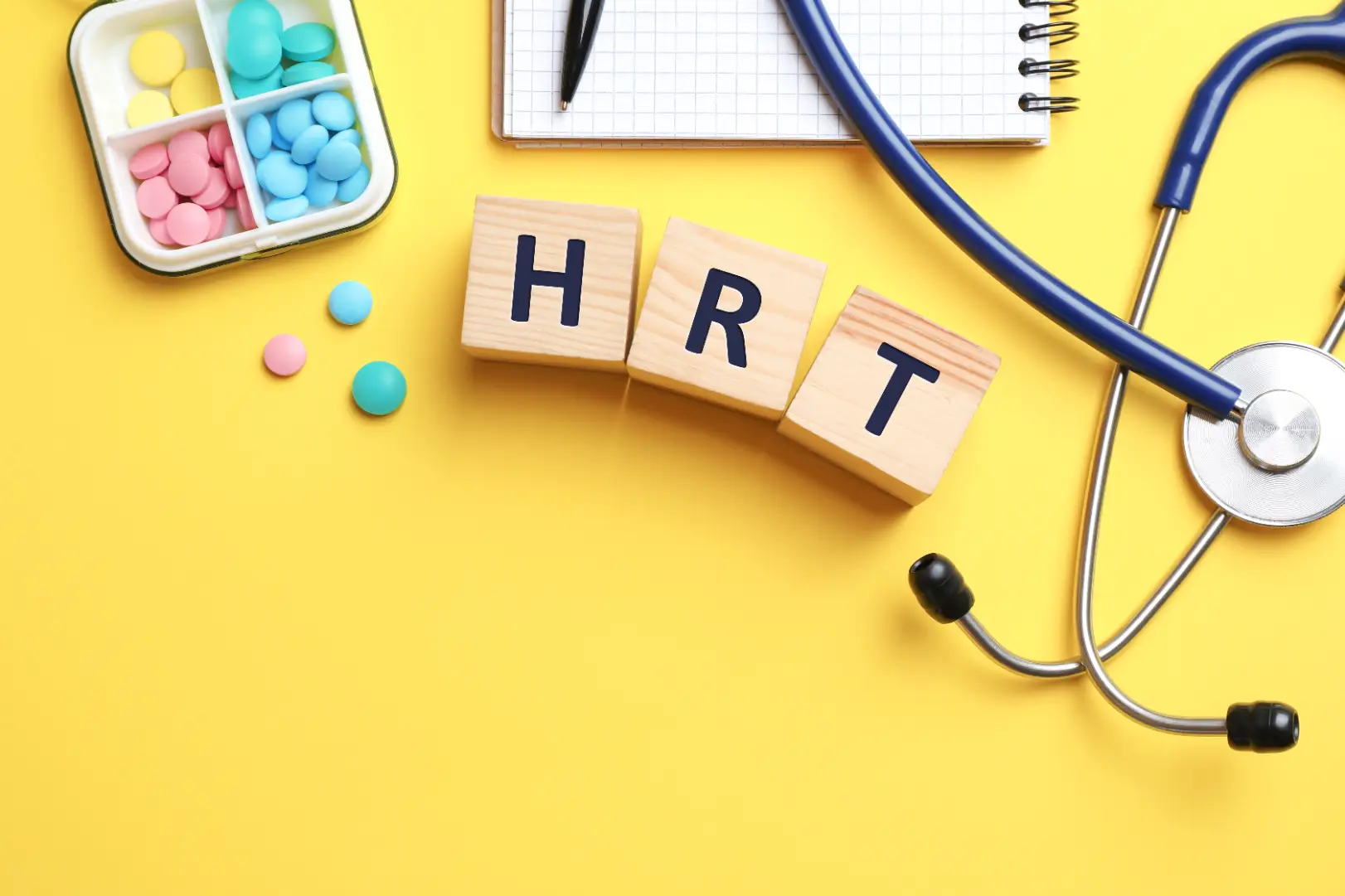 Wooden blocks spelling HRT beside colorful tablets, a stethoscope, notebook, and pen on a yellow surface.